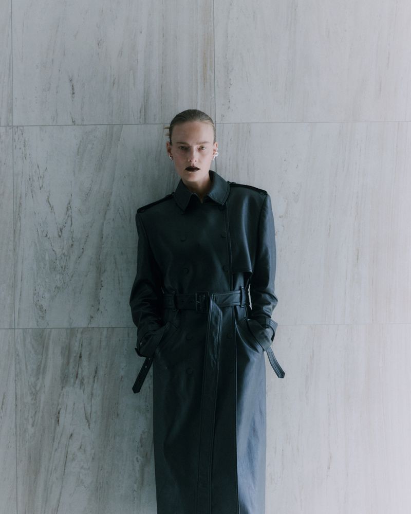 Model Ania Kisiel posed in a long black belted leather trench coat against a pale marble wall.