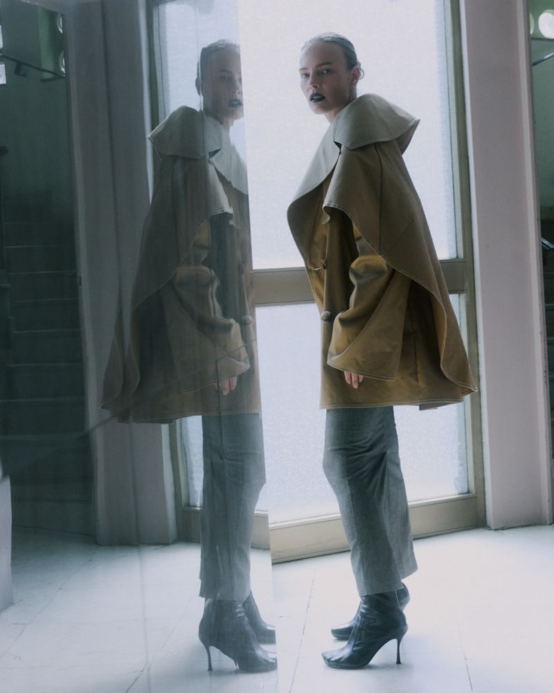 Indoor fashion shot of an adult female model in a tan coat and grey pants, standing near a bright window with her reflection visible.