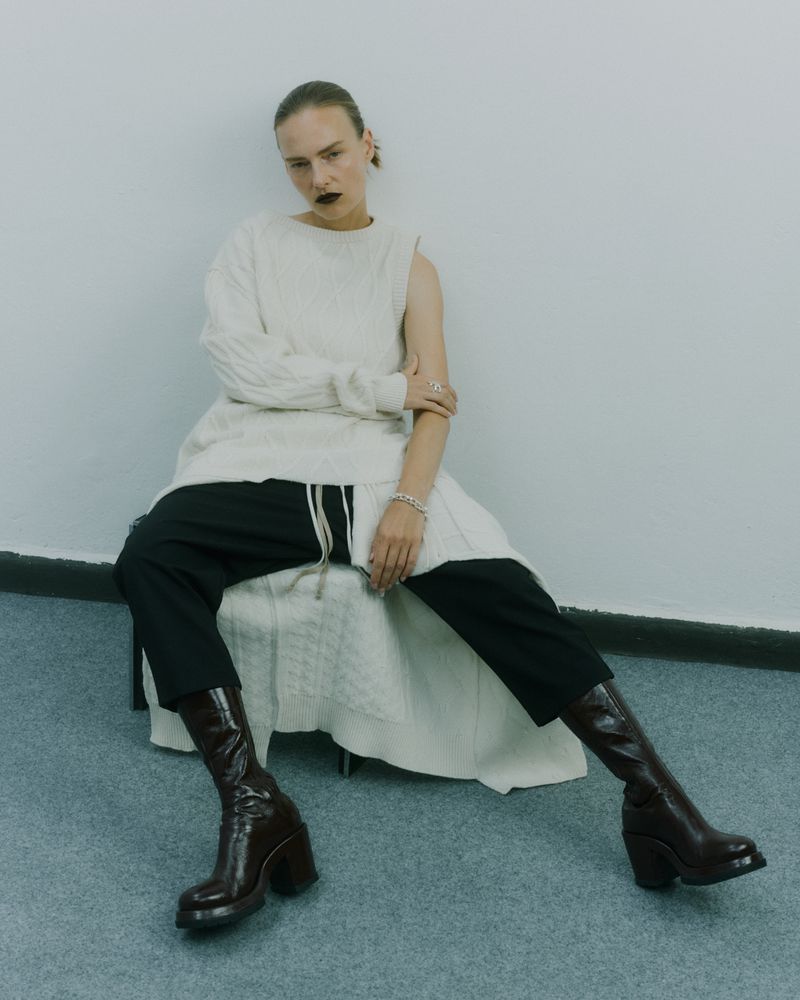 Fashion editorial shot of a woman in a white knit dress, black pants, and brown leather boots, seated in a simple indoor studio setting.