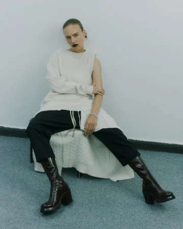 Fashion editorial shot of a woman in a white knit dress, black pants, and brown leather boots, seated in a simple indoor studio setting.
