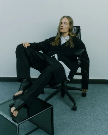Model Ania Kisiel lounges in a black office chair wearing an oversized sweater and trousers with feet resting on a glass table.