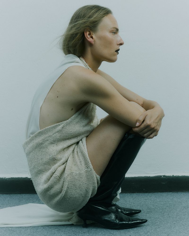 Portrait of an adult female crouching indoors, wearing a light-colored dress, knee-high boots, and dark lipstick against a white background.
