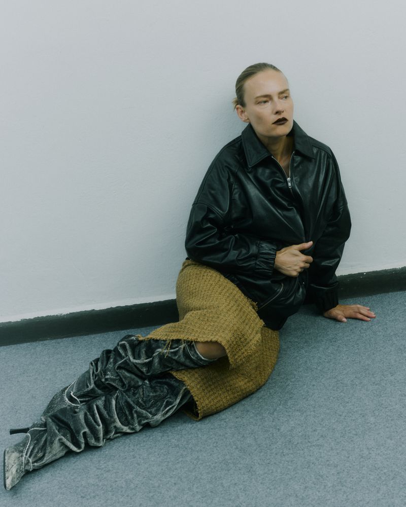 Adult woman poses in a black jacket and brown skirt, seated on a grey floor against a white wall with grey heeled velvet boots.