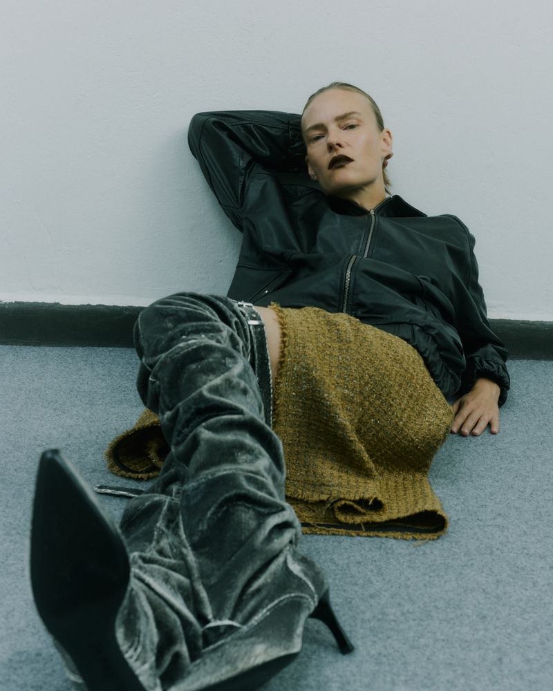 Woman in a black bomber jacket and gold skirt poses while sitting on the floor wearing grey high-heeled boots.