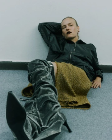 Woman in a black bomber jacket and gold skirt poses while sitting on the floor wearing grey high-heeled boots.