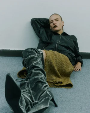 Model Ania Kisiel reclines against a wall wearing a black zip jacket, mustard skirt, and slouchy velvet thigh-high boots.