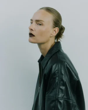 Model Ania Kisiel in an oversized black leather jacket with dark lipstick posing against a pale studio background.