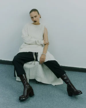 Model Ania Kisiel seated wearing an off-white asymmetrical knit top, black trousers, and dark burgundy leather boots.