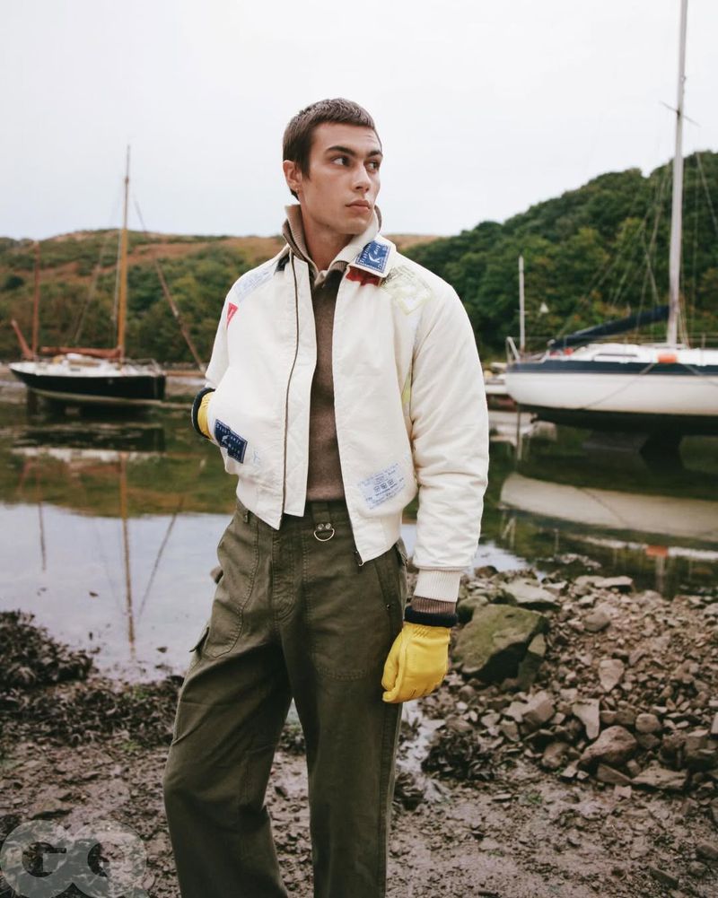 A young adult man in a bomber jacket poses on a rocky beach with boats in the background, creating a fashion editorial shot.