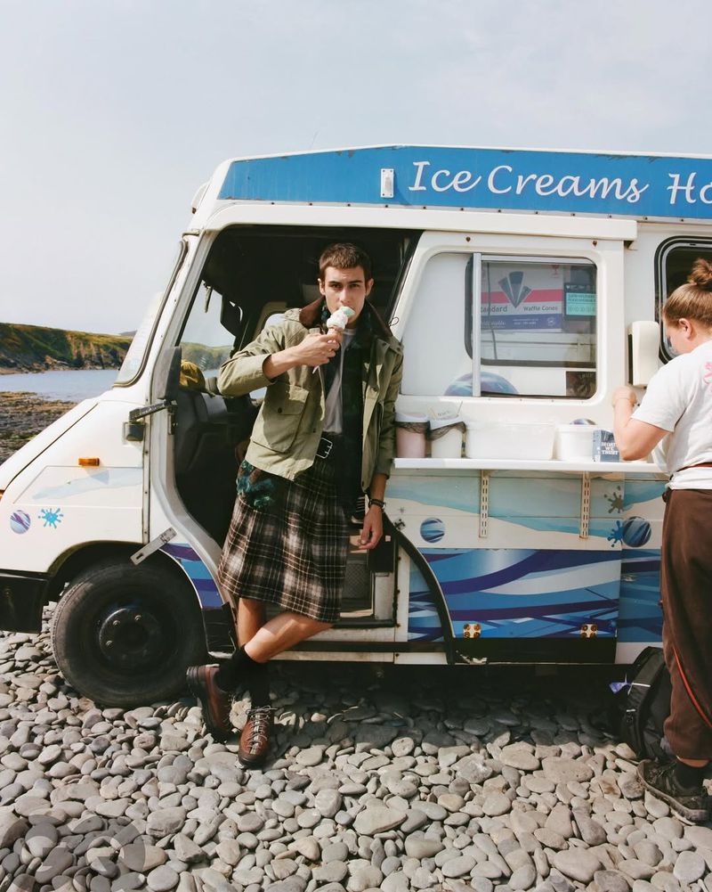 A man in a kilt enjoys ice cream in front of an ice cream truck on a rocky shore, offering a unique and playful visual narrative.