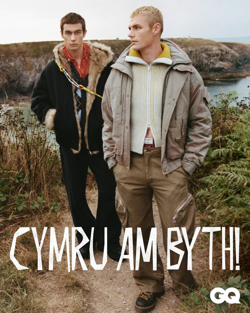 Two men pose on a path near a cliff and the sea, wearing fashionable jackets and trousers in an outdoor fashion editorial.