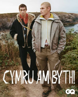 Two male models standing on a dirt path outdoors near a body of water, wearing winter outerwear.