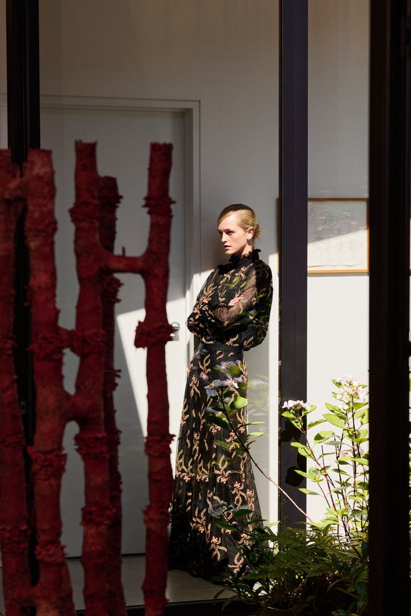 A fair-skinned woman with short blonde hair poses indoors in a black floral gown, with a red sculpture and potted plants nearby.