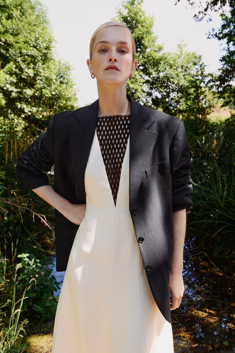 A fair-skinned woman in a black blazer and cream-colored dress poses in an outdoor setting with a blurred green background.