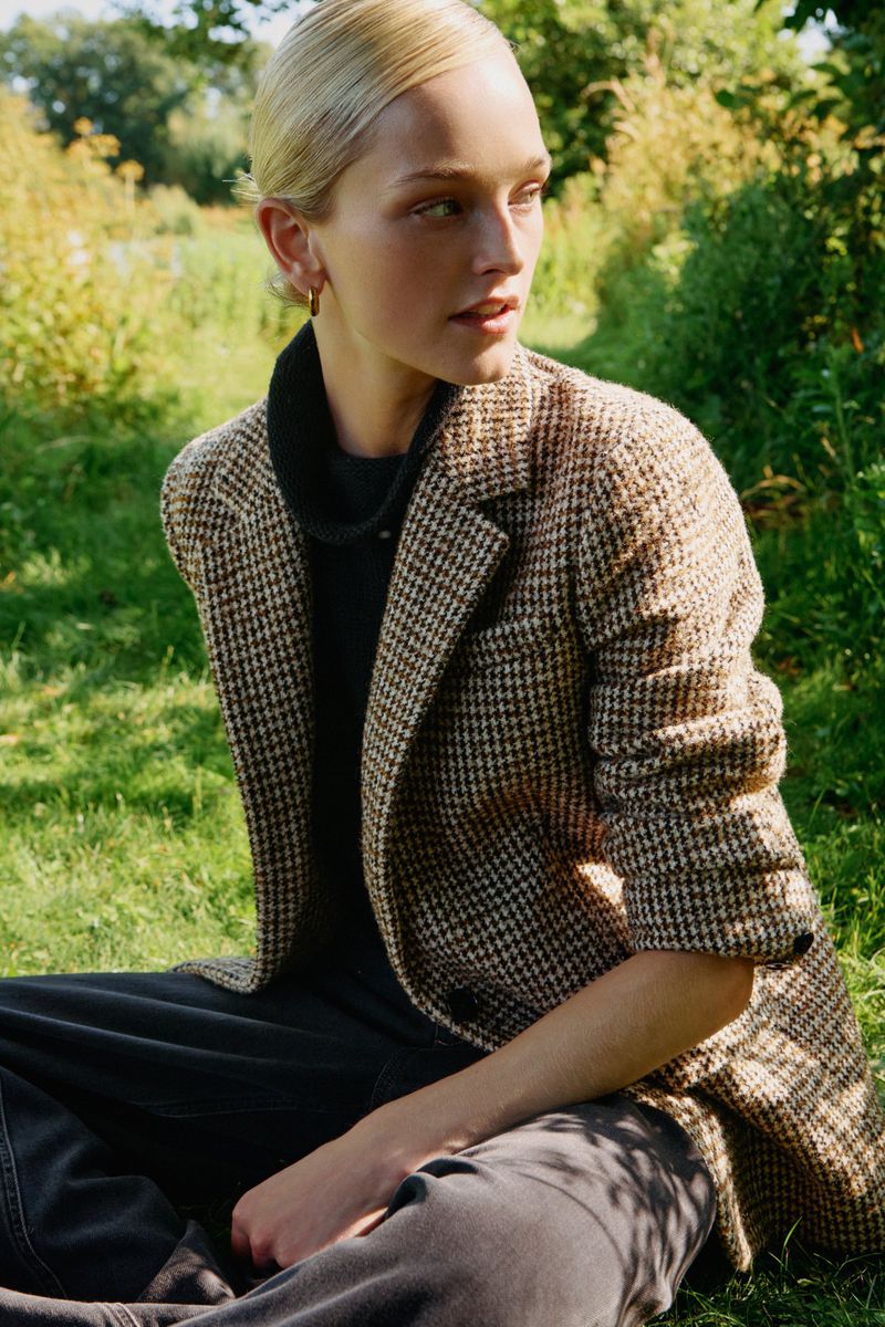 A fair-skinned woman with blonde hair poses outdoors in a houndstooth blazer, black top, and jeans, seated on green grass with a blurred background.