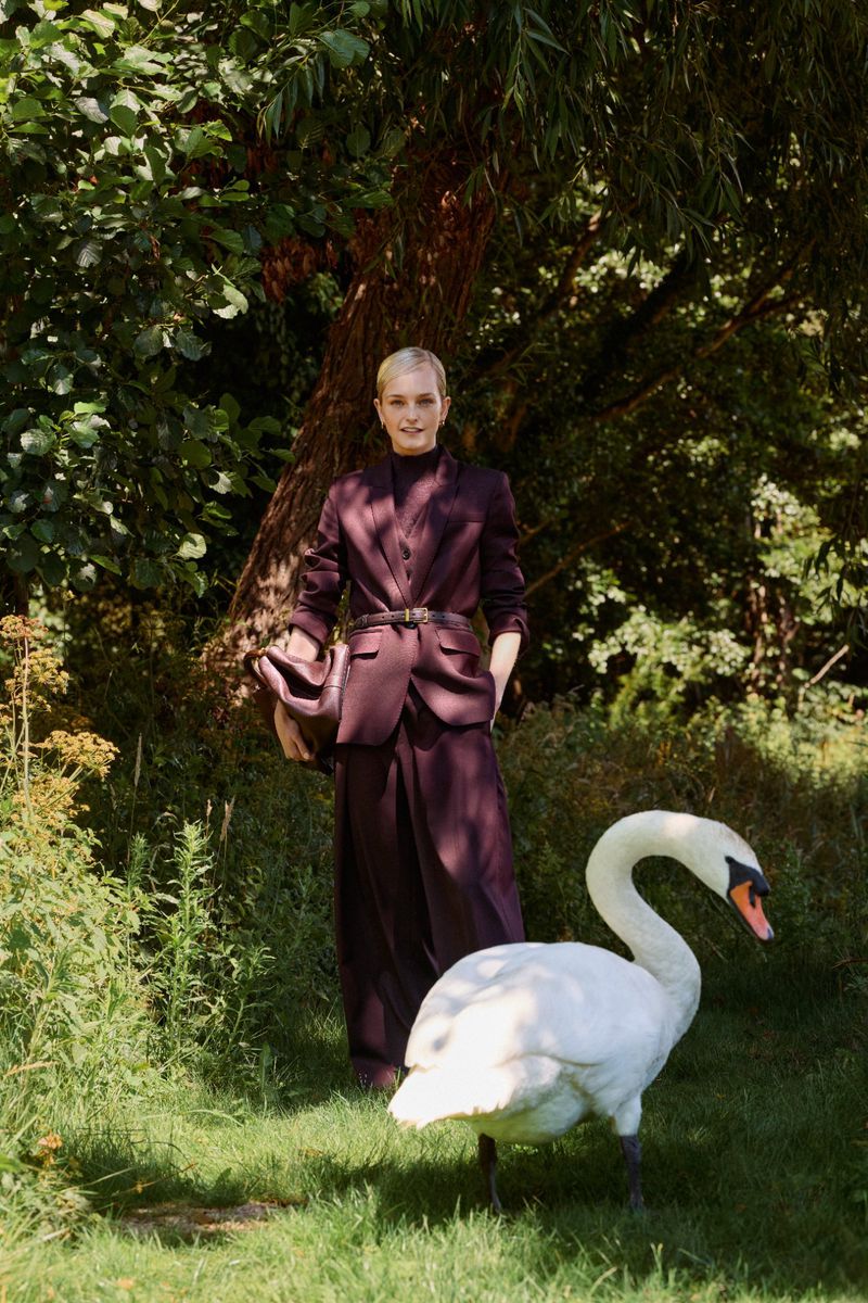 Woman in a burgundy suit poses in a sun-dappled glade with a swan standing in front of her in a striking outdoor fashion portrait.