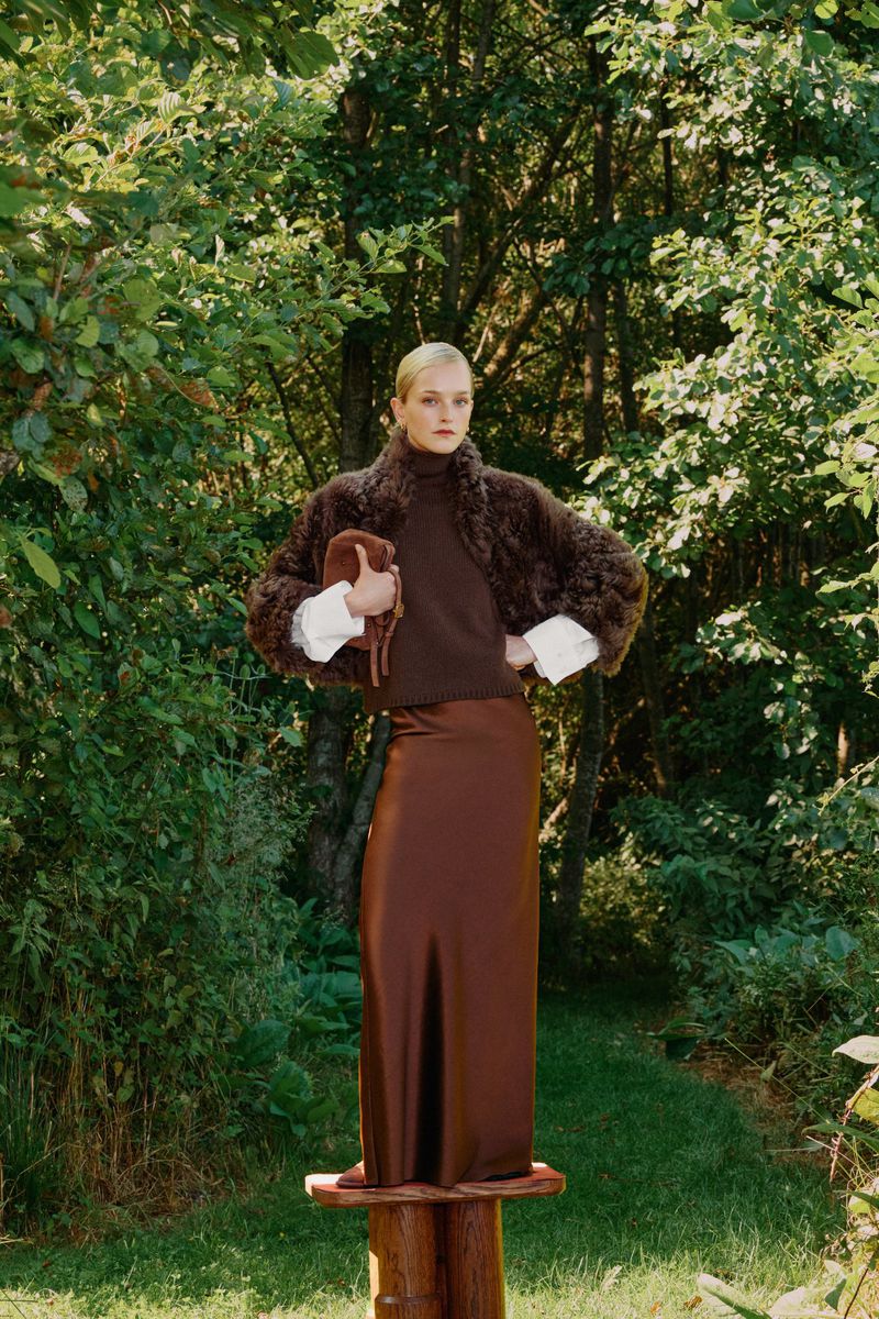 Elegant woman in brown turtleneck, fur-like jacket, and satin maxi skirt poses on a wooden stool outdoors surrounded by lush greenery.