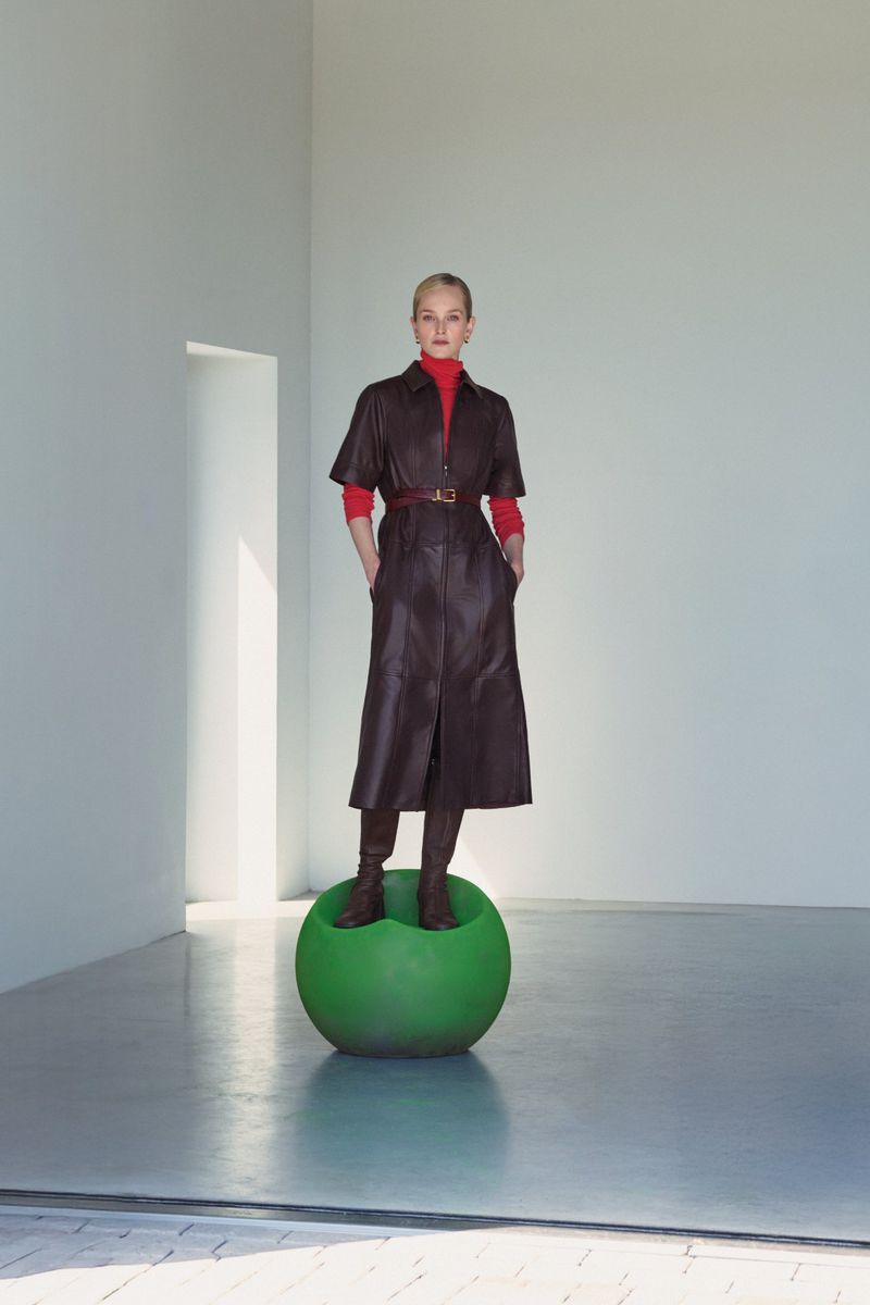 Fashion shot of a blonde woman in a brown leather dress, red top, and brown boots standing on a green sphere in a white corner space.