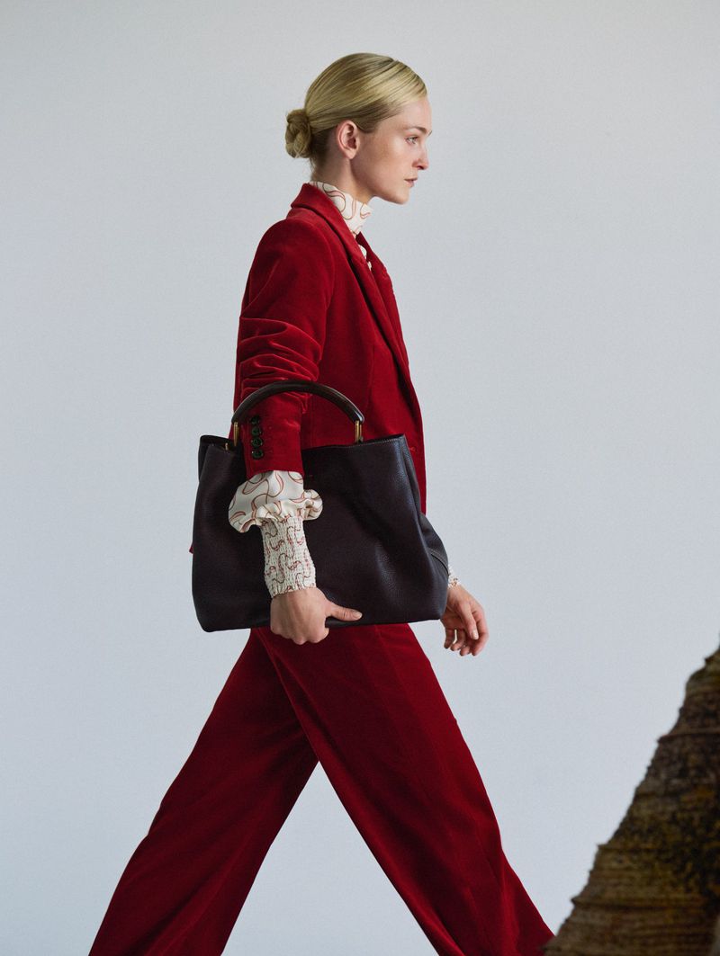 A woman in a red velvet suit walks, carrying a black tote bag. She has her blonde hair in a bun, posing against a light gray background.