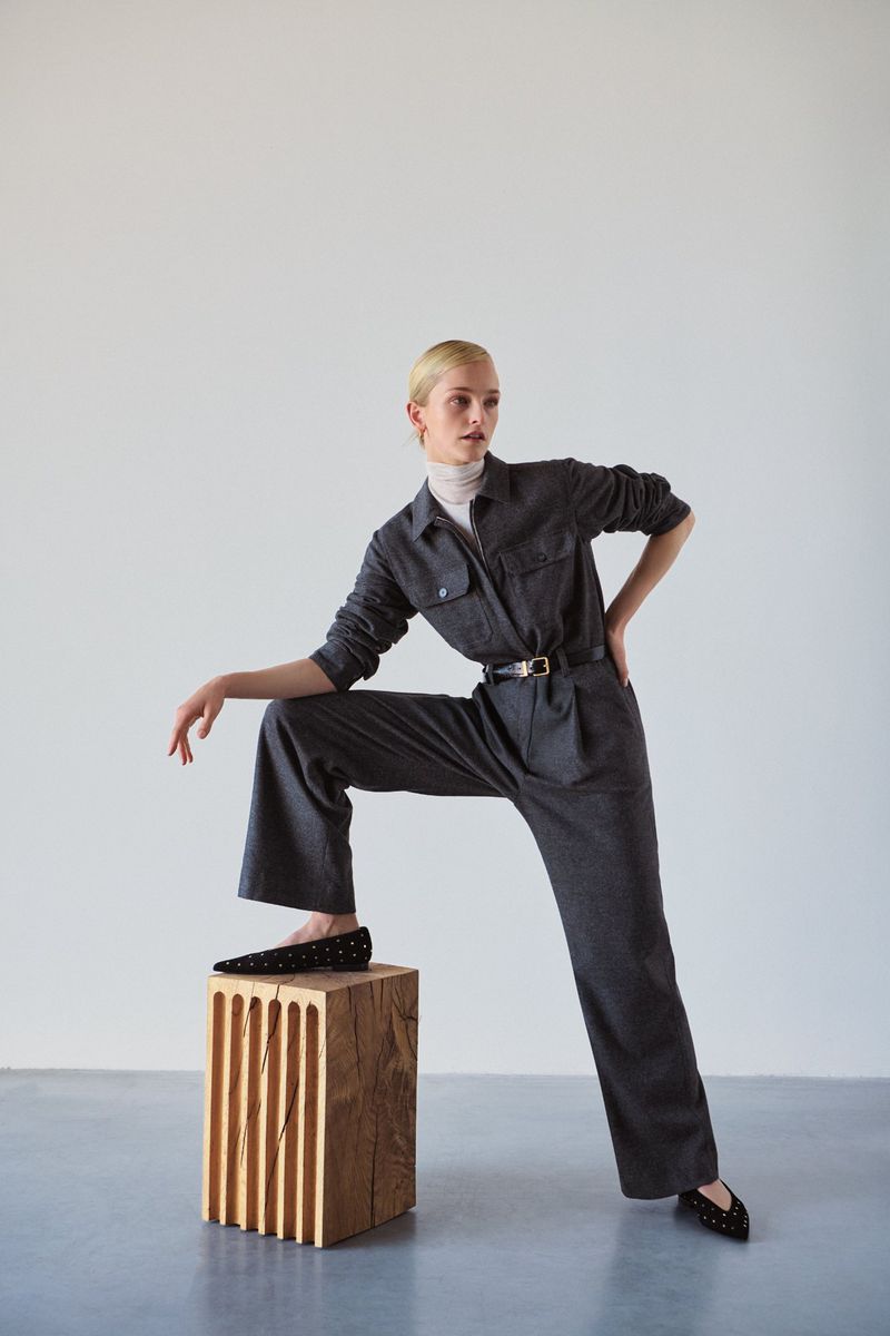 Fashion shot of a woman in a grey jumpsuit standing with one foot on a wooden block, posing against a neutral background in a studio.