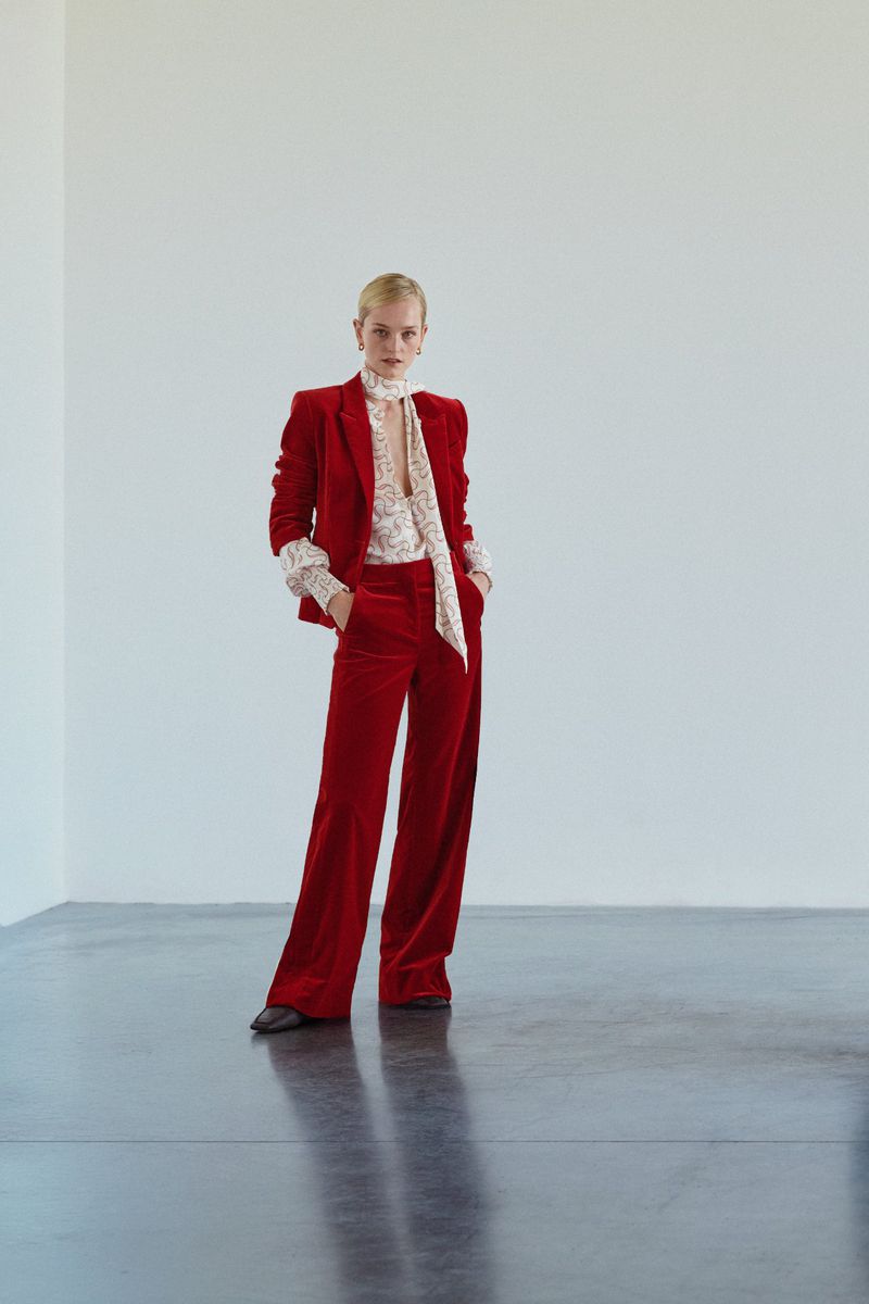 Woman poses in a red velvet suit with a floral blouse against a white background in a studio setting, fashion portrait.