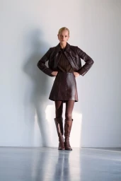 Jean Campbell wearing a brown leather jacket, matching mini skirt, and knee-high boots against a white studio wall.