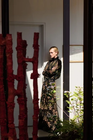 Model Jean Campbell wearing a black embroidered maxi dress standing in a sunlit room beside a red sculpture for ME+EM.