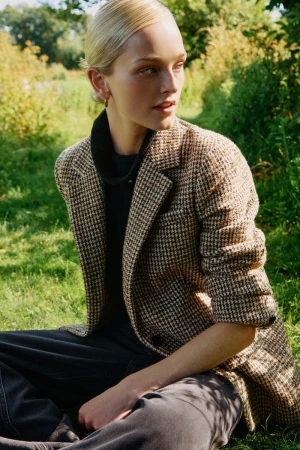 Model Jean Campbell seated outdoors in grass wearing a brown houndstooth blazer and dark turtleneck.