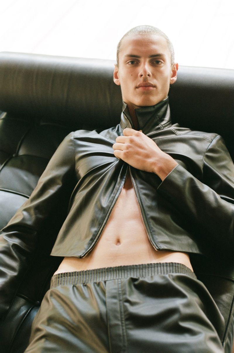 A fair-skinned male model with a shaved head, wearing an open black leather jacket, poses on a black leather couch in a studio.