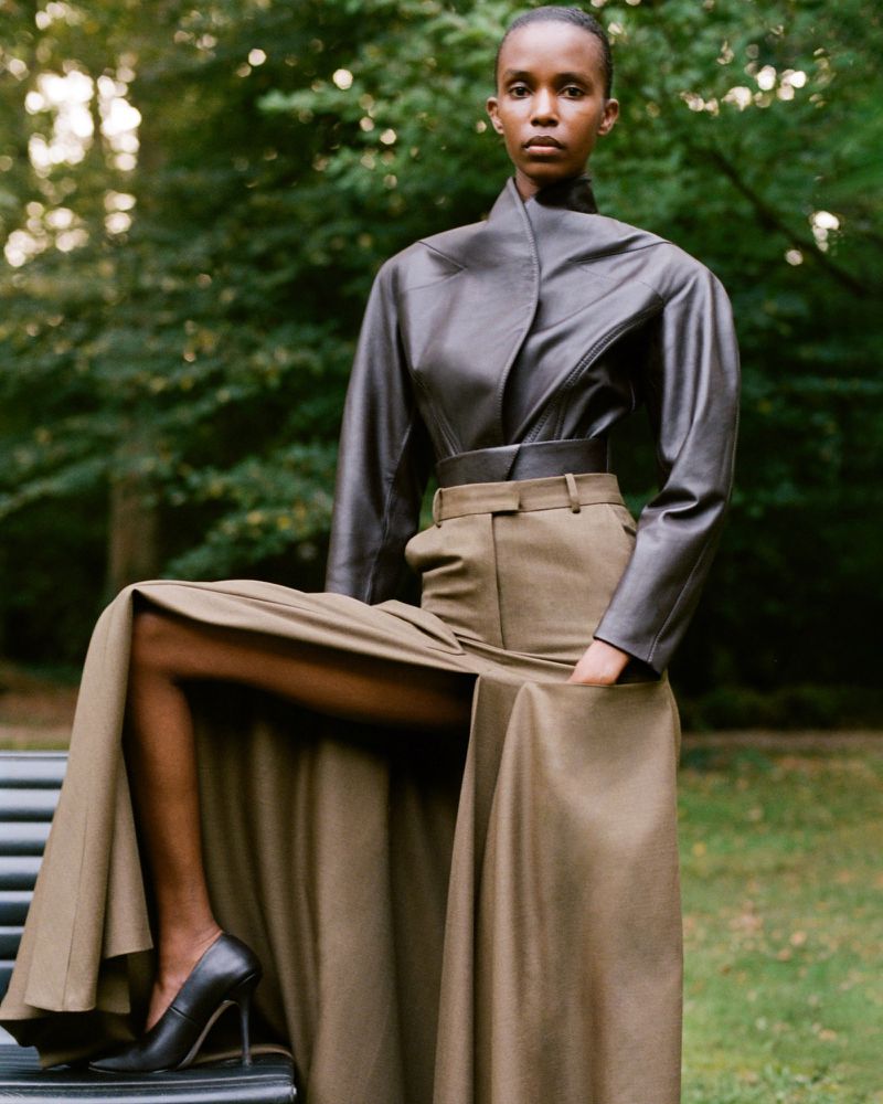 An adult female wearing a leather jacket, olive high-waisted pants, and black high heels poses on a park bench in an outdoor setting.
