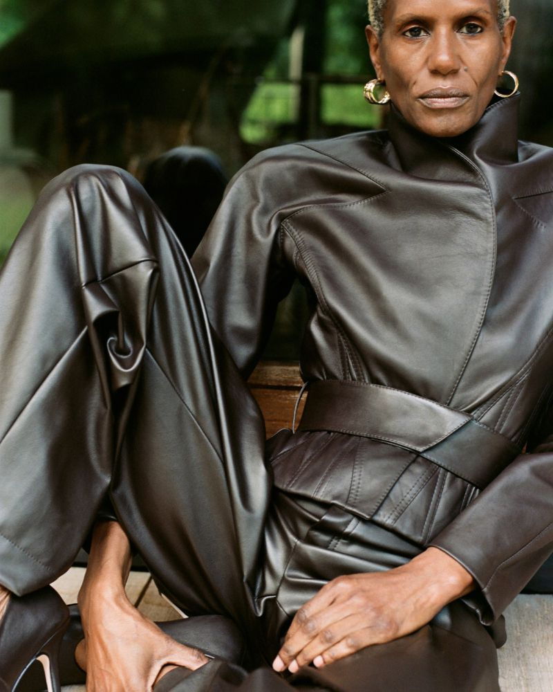 Adult female with short grey hair poses in a brown leather jumpsuit and black high heels sitting outdoors with a lush green background.