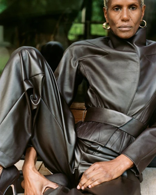 Adult female with short grey hair poses in a brown leather jumpsuit and black high heels sitting outdoors with a lush green background.