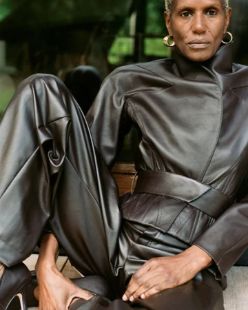 Adult female with short grey hair poses in a brown leather jumpsuit and black high heels sitting outdoors with a lush green background.