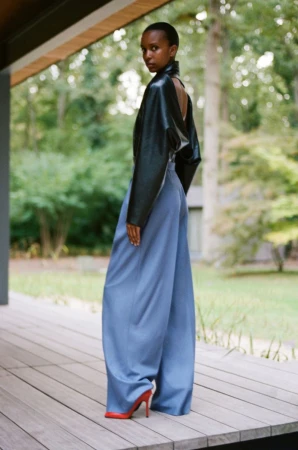 Model wearing a black open-back leather jacket, oversized blue wide-leg trousers, and red heels on a wooden deck.