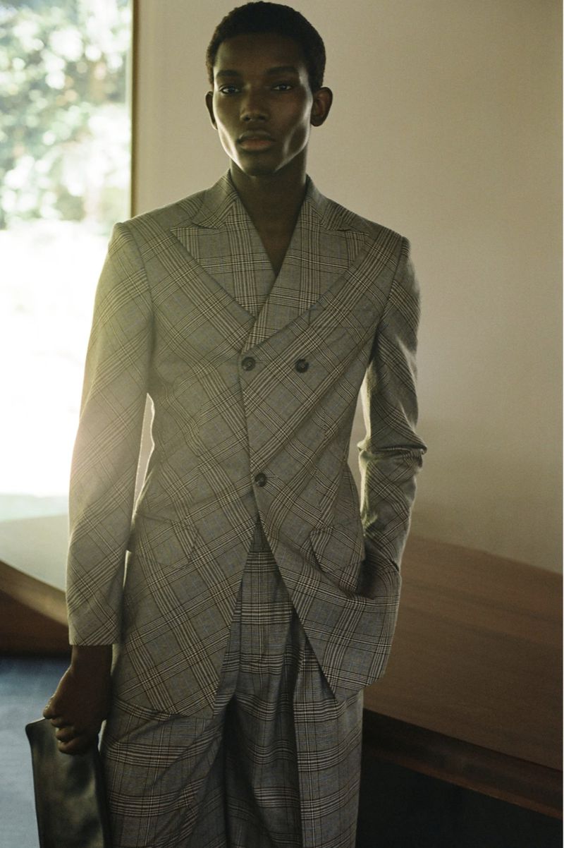 A dark-skinned man in a grey plaid suit poses indoors, holding a black bag, showcasing a modern and sophisticated fashion aesthetic.