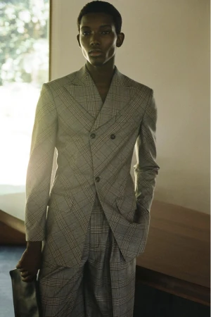 A dark-skinned man in a grey plaid suit poses indoors, holding a black bag, showcasing a modern and sophisticated fashion aesthetic.