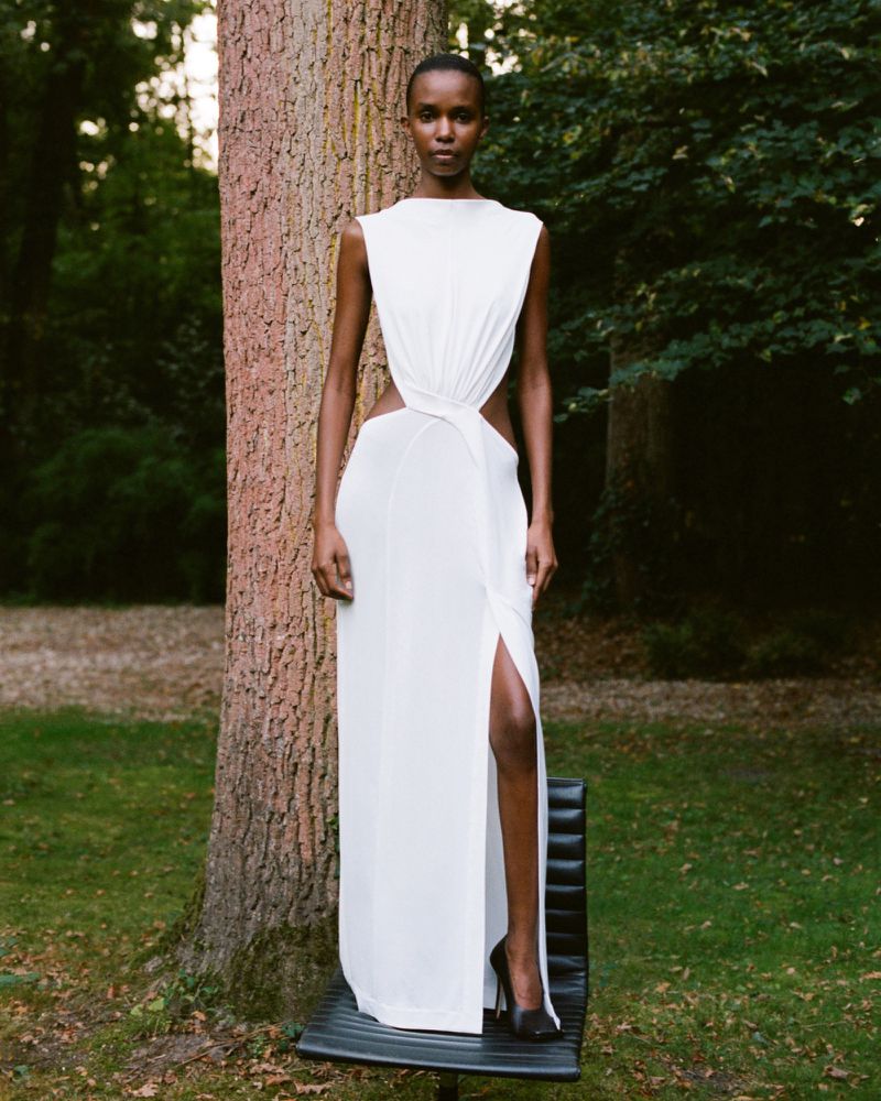 Woman in elegant white gown standing on a chair outdoors. Fashionable dress with cut-out design, tree in the background, outdoor setting.