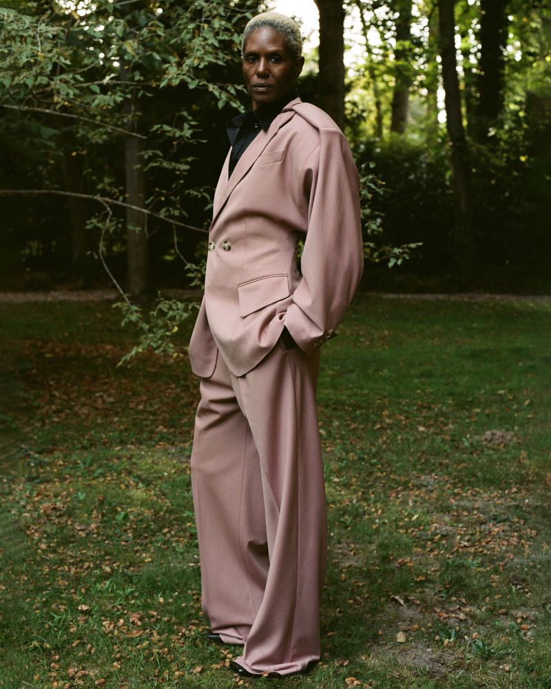 Model posing in an oversized dusty rose tailored suit and black collared shirt on a grassy lawn backed by lush trees.