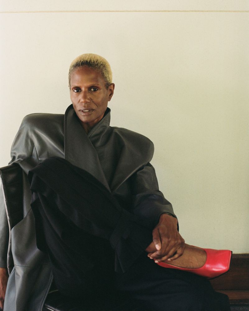A woman with short blonde hair sits, wearing a grey oversized coat and black pants, with a red flat shoe visible, in a studio setting.