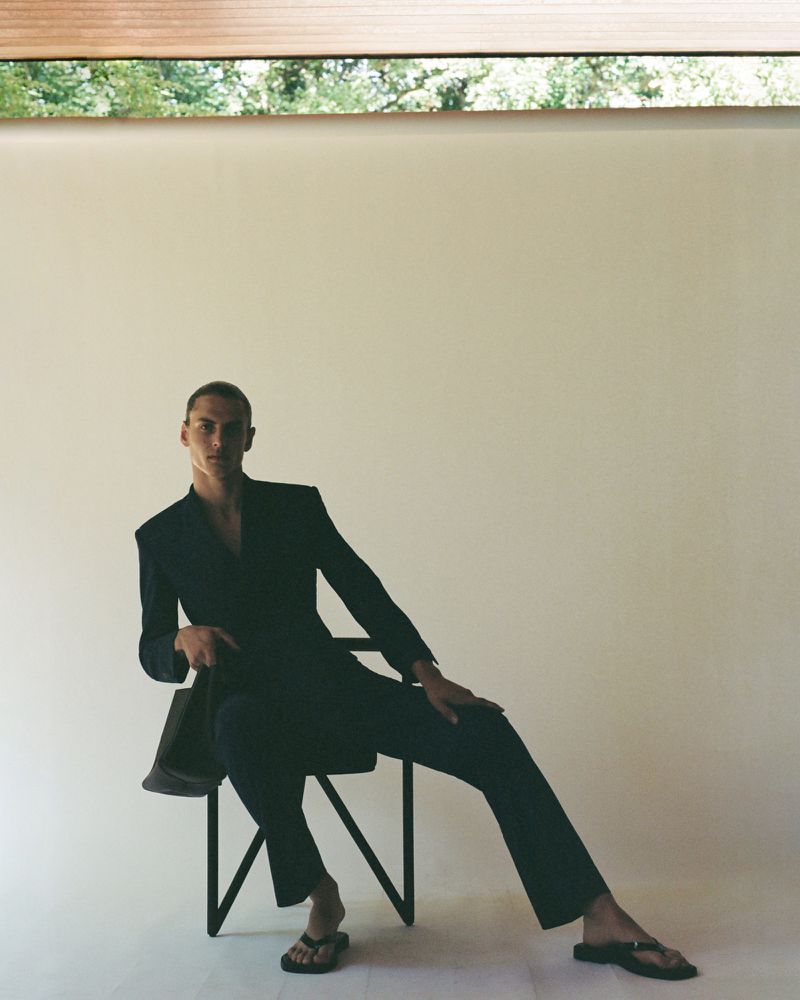 A man seated on a chair wearing a black tailored blazer, matching trousers, and flip-flops against a neutral studio backdrop.
