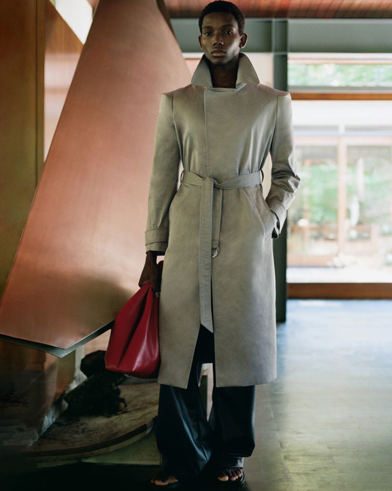 An adult male poses indoors wearing a light grey trench coat, dark pants, and carrying a red bag, with a modern geometric background.