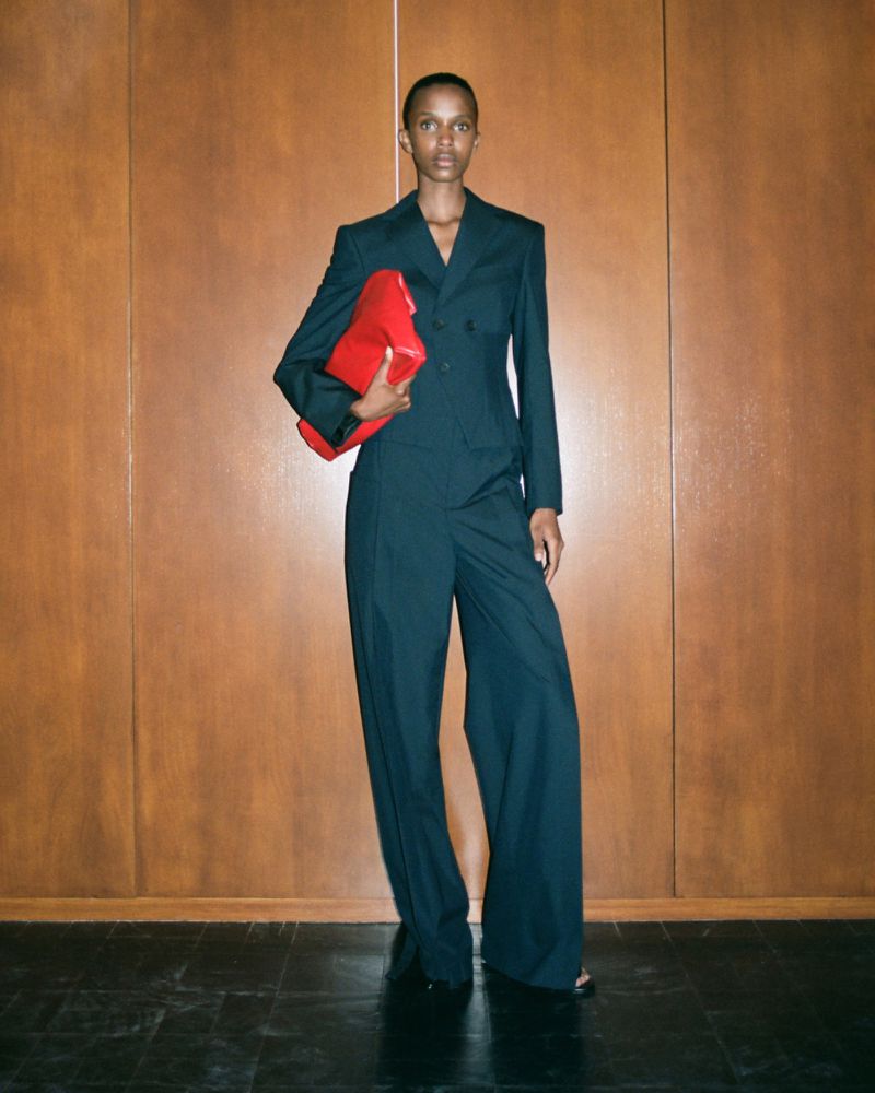 A tall, Black woman stands in a formal suit against a wood paneled wall, holding a bright red clutch in a fashion forward pose.