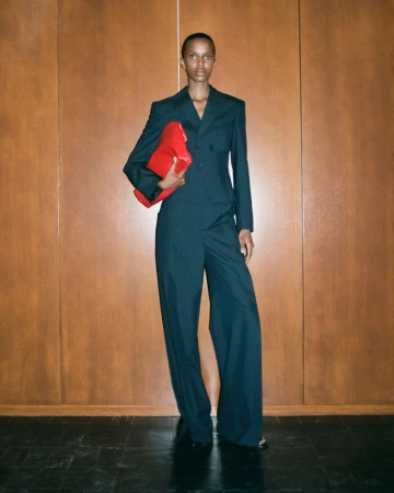 A tall, Black woman stands in a formal suit against a wood paneled wall, holding a bright red clutch in a fashion forward pose.