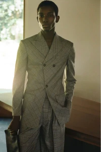 A Black male model wearing a grey plaid double-breasted suit with wide lapels standing near an indoor window with soft lighting.