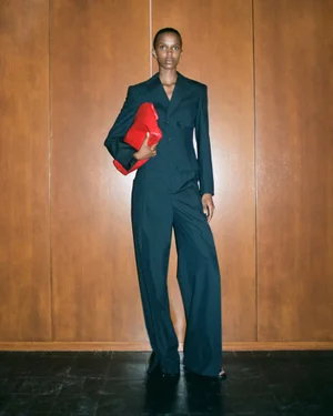 Model standing against a wood-paneled wall wearing a dark teal tailored suit and holding a vibrant red leather clutch.