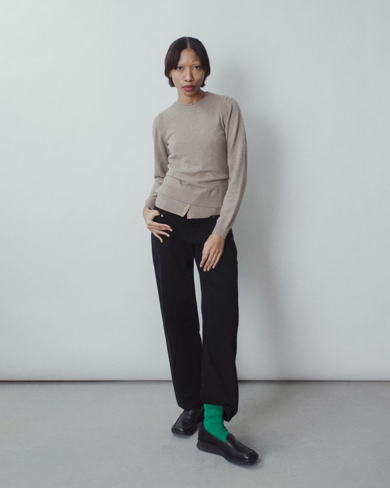 Full-length portrait of an Asian female model in a taupe sweater, black pants, and green socks in a studio setting, posing fashionably.