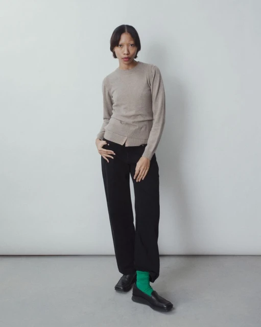 Full-length portrait of an Asian female model in a taupe sweater, black pants, and green socks in a studio setting, posing fashionably.