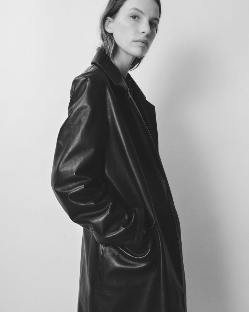 Indoor shot of a woman in a black faux-leather trench coat standing against a light gray wall in a fashion editorial style.