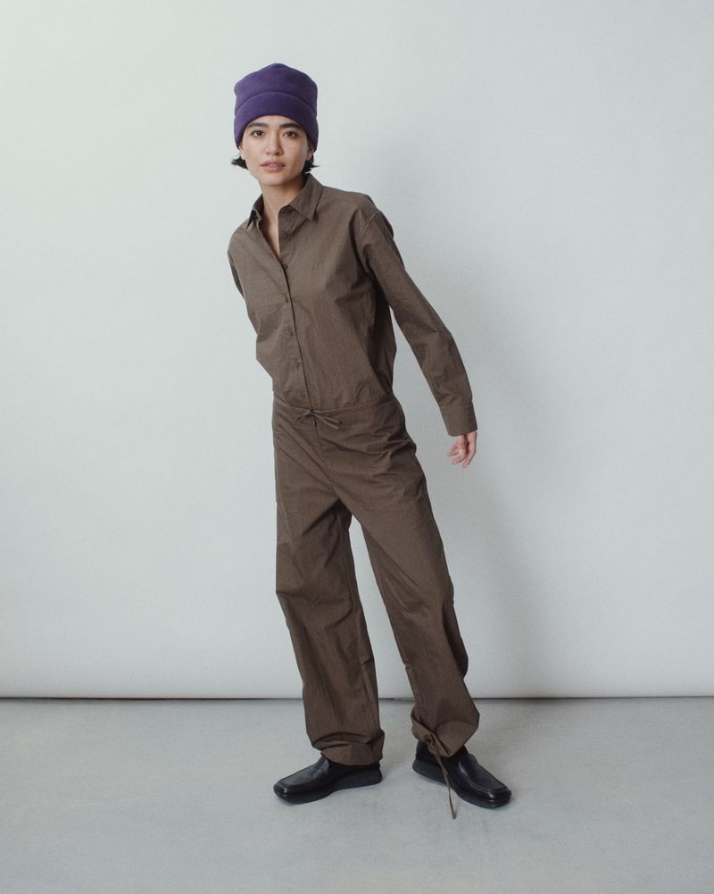 Adult female in brown coveralls and a purple knit hat strikes a full pose in a brightly lit photo studio against a neutral backdrop.