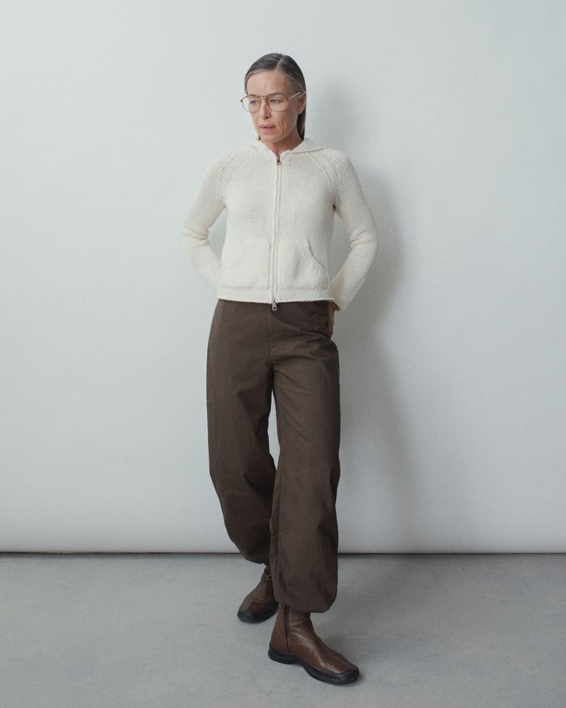 Full-length shot of an adult female with glasses, wearing a cream-colored knitted jacket, loose brown pants, and brown boots against a neutral wall.